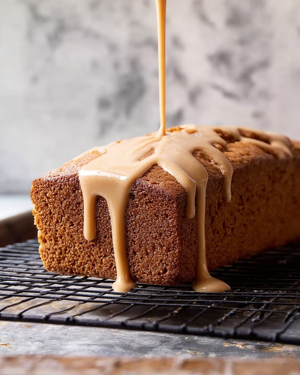 A single rectangular loaf with a brown, slightly rough and cooked outer crust rests on a black wire cooling rack over a white marbled surface. A light beige, smooth glaze is being poured over the top right corner of the loaf, dripping down the side in thick streams. The glaze is shiny and contrasts with the darker texture of the loaf underneath. The background is softly blurred with warm colors. Photo taken with an iphone --ar 4:5 --v 7