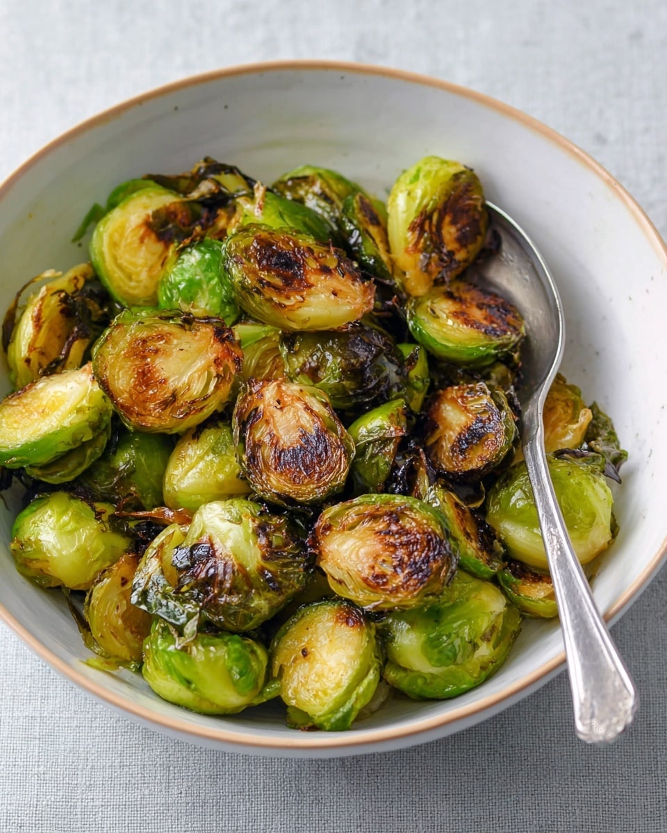 A white bowl filled with cooked Brussels sprouts showing about two layers. The bottom layer has bright green Brussels sprouts with a smooth texture. The top layer has Brussels sprouts with a mix of bright green and browned parts, some crispy and slightly darkened edges. A silver spoon rests inside the bowl on the right side. The bowl sits on a white marbled surface. photo taken with an iphone --ar 4:5 --v 7