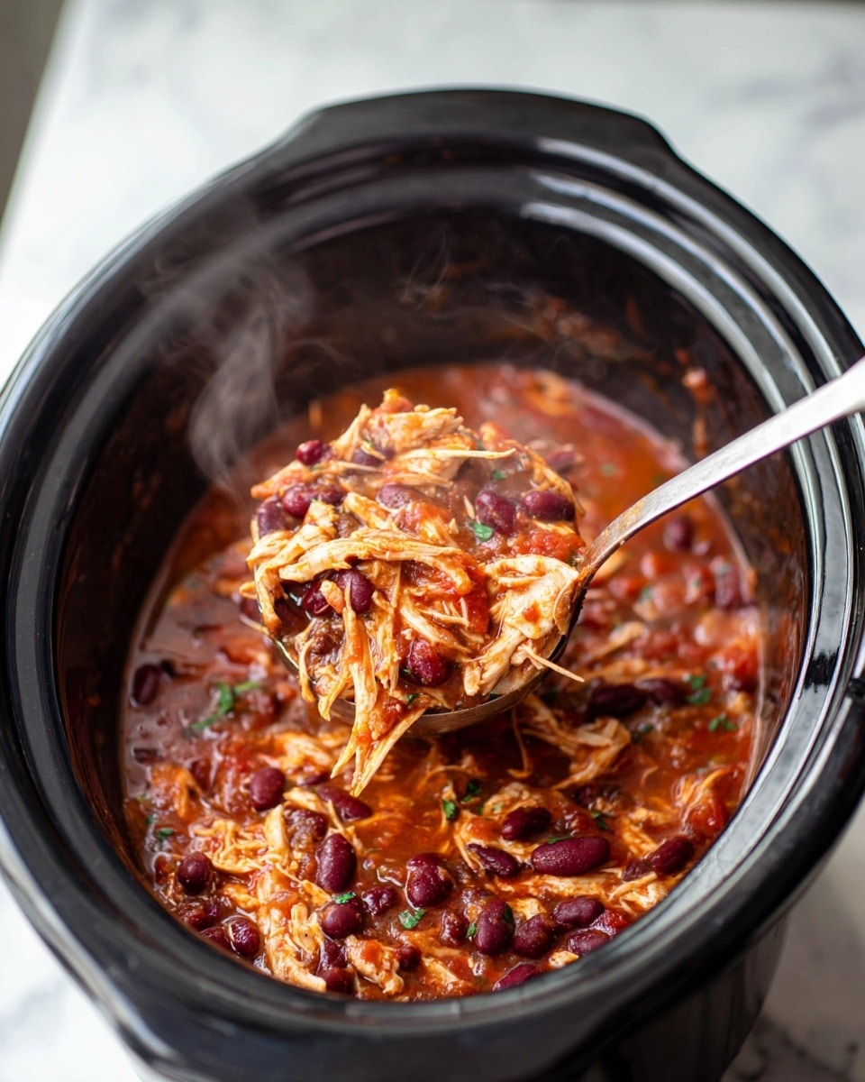 A close-up view of a slow cooker filled with a chunky stew showing shredded white chicken mixed with dark red kidney beans and small pieces of green vegetables in a thick red tomato sauce. The stew looks hot with steam rising from it. A large metal ladle with a shiny handle is lifting a portion of the stew above the slow cooker’s black ceramic pot. The background has a white marbled texture. photo taken with an iphone --ar 4:5 --v 7