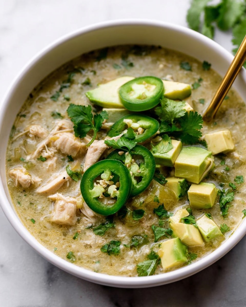 A white bowl filled with a thick greenish soup made of shredded pieces of chicken and small bits of herbs, topped with fresh slices of jalapeño peppers, bright green avocado slices arranged neatly, and sprigs of fresh cilantro. A gold spoon is partially submerged inside the soup toward the front of the bowl, resting on the edge. The bowl is placed on a white marbled surface. Photo taken with an iphone --ar 4:5 --v 7