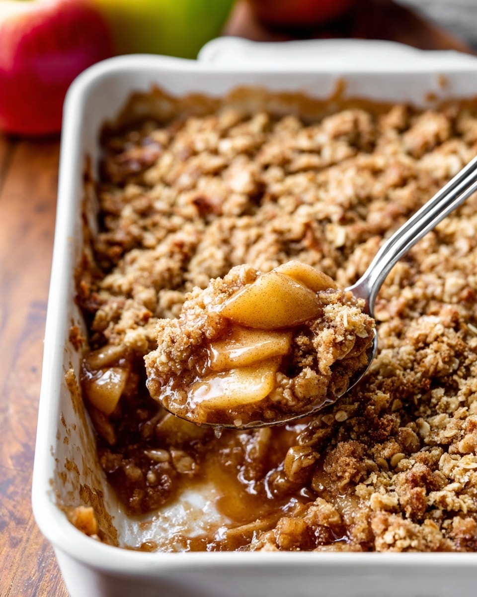 A white baking dish holds a warm apple crumble with two visible layers: the bottom layer is light golden, chunky apple pieces mixed with a syrupy sauce, and the top layer is a crumbly, textured oat and brown sugar topping that looks toasted and crisp. A spoon inside the dish scoops out some of the apple filling from the corner. The baking dish is placed on a wooden surface, with blurred apples in the foreground. photo taken with an iphone --ar 4:5 --v 7