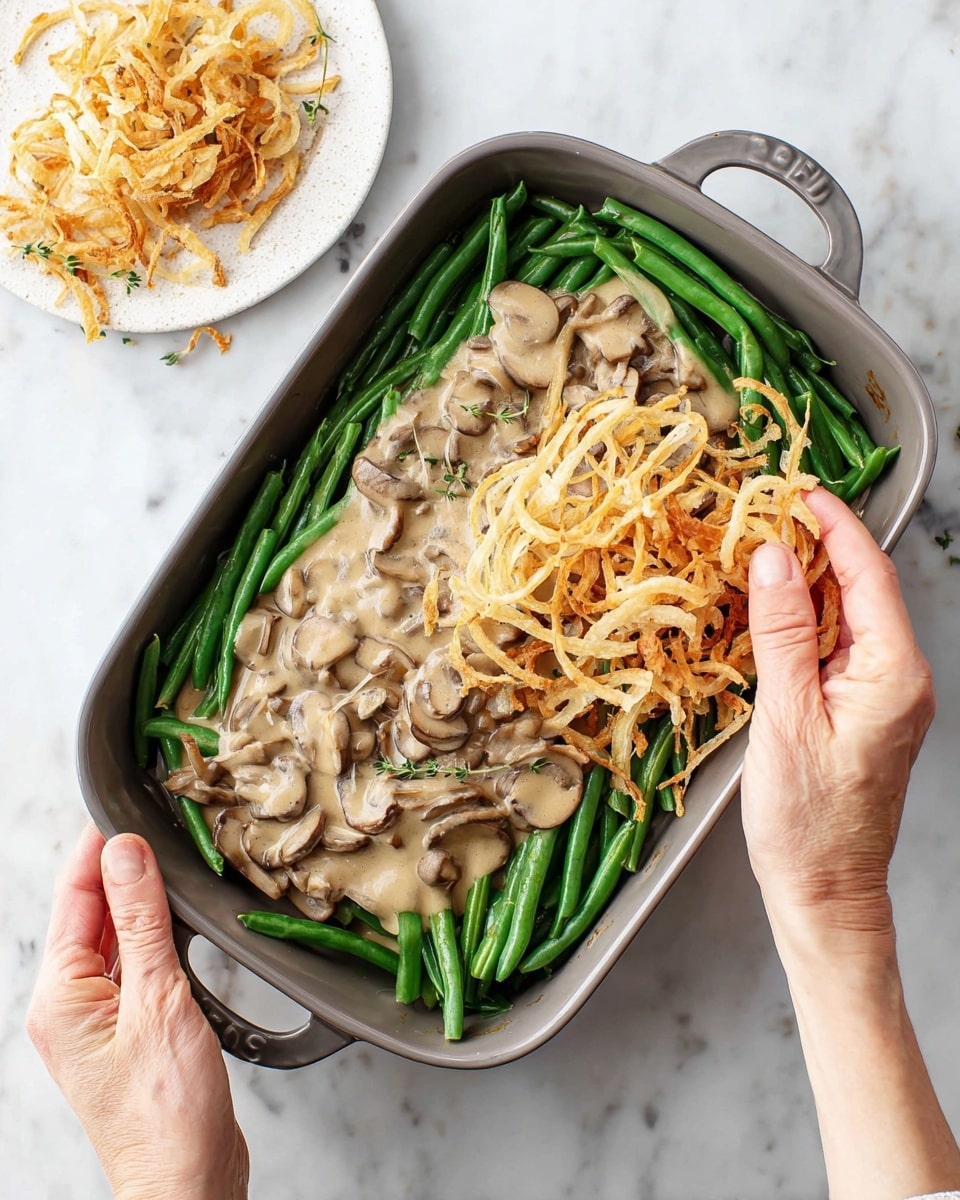 A rectangular gray baking dish on a white marbled surface holds neatly arranged green beans forming the bottom layer. Above the beans, there is a thick light brown mushroom sauce with visible mushroom slices covering the center. A woman's hands are adding a top layer of thin, crispy golden onion strings, some of which are still on a small round white plate to the side. The contrast between the fresh green beans, creamy sauce, and crunchy onion topping is clear and appealing. Photo taken with an iphone --ar 4:5 --v 7