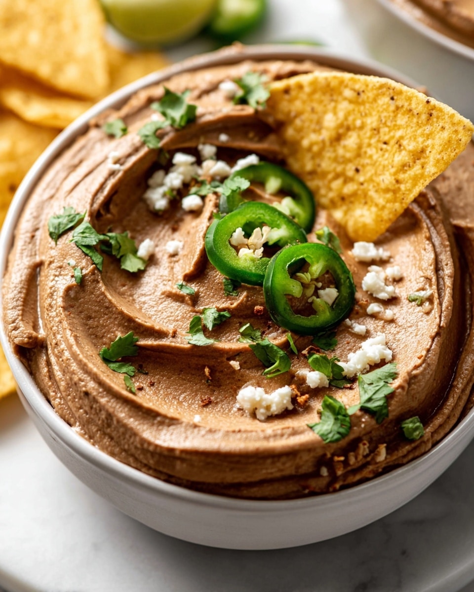 A close-up of a smooth, creamy brown dip swirled evenly inside a white bowl, topped with small scattered white cheese crumbles and fresh cilantro leaves. A few slices of bright green jalapeño peppers are placed on top, adding a pop of color. A crisp yellow tortilla chip is dipped into the smooth texture of the dip, partially covered. The bowl sits on a brown wooden surface, with a clean white marbled texture in the background. Photo taken with an iphone --ar 4:5 --v 7