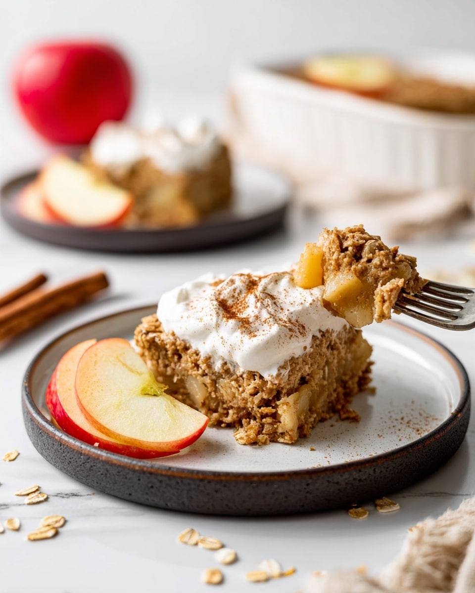 The image shows a close-up of a slice of apple oat dessert on a round white plate with a dark rim, placed on a white marbled surface. The dessert slice has two distinct layers: a dense, textured bottom layer filled with chunks of apple and oats in golden brown tones, and a topping of whipped cream sprinkled with cinnamon, giving it a soft white and light brown look. Next to the dessert are two fresh apple slices with shiny red and yellow skin. A fork holds a bite of the dessert with whipped cream. In the background, there is a whole red apple and another slice of the dessert on a white plate with a dark rim, blurred out. Scattered bits of oats and cinnamon sticks add rustic charm to the scene. Photo taken with an iphone --ar 4:5 --v 7