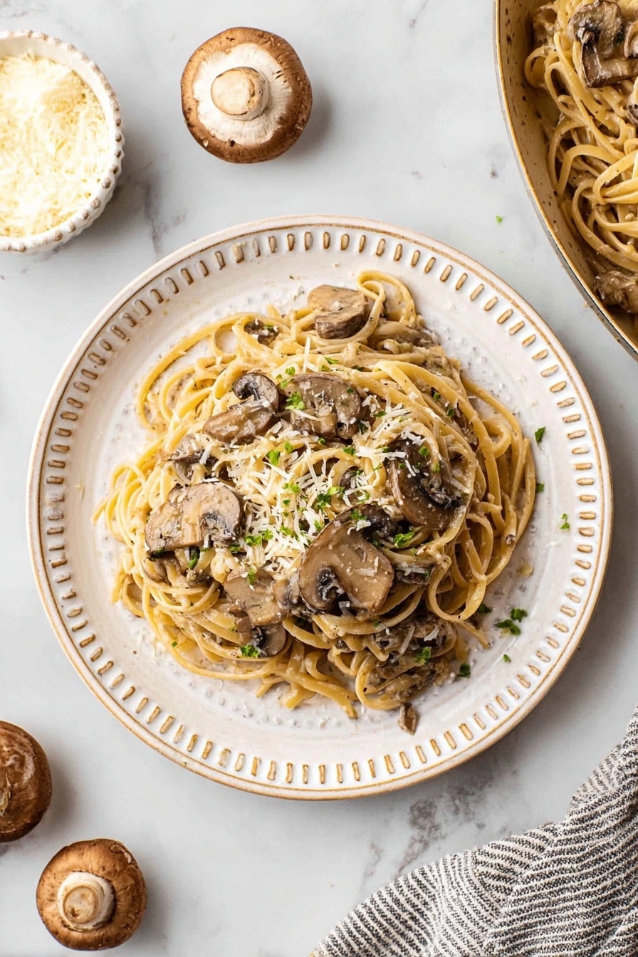 A white plate with a bumpy rim holds a serving of creamy mushroom pasta. The pasta is flat and light brown, mixed evenly with dark sliced mushrooms covered in a light cream sauce. On top, small green parsley bits and white cheese flakes add texture and color. The plate sits on a white marbled surface, with some whole mushrooms and a small white bowl of cheese nearby. A striped cloth is partially visible under the pan of more pasta. Photo taken with an iphone --ar 4:5 --v 7