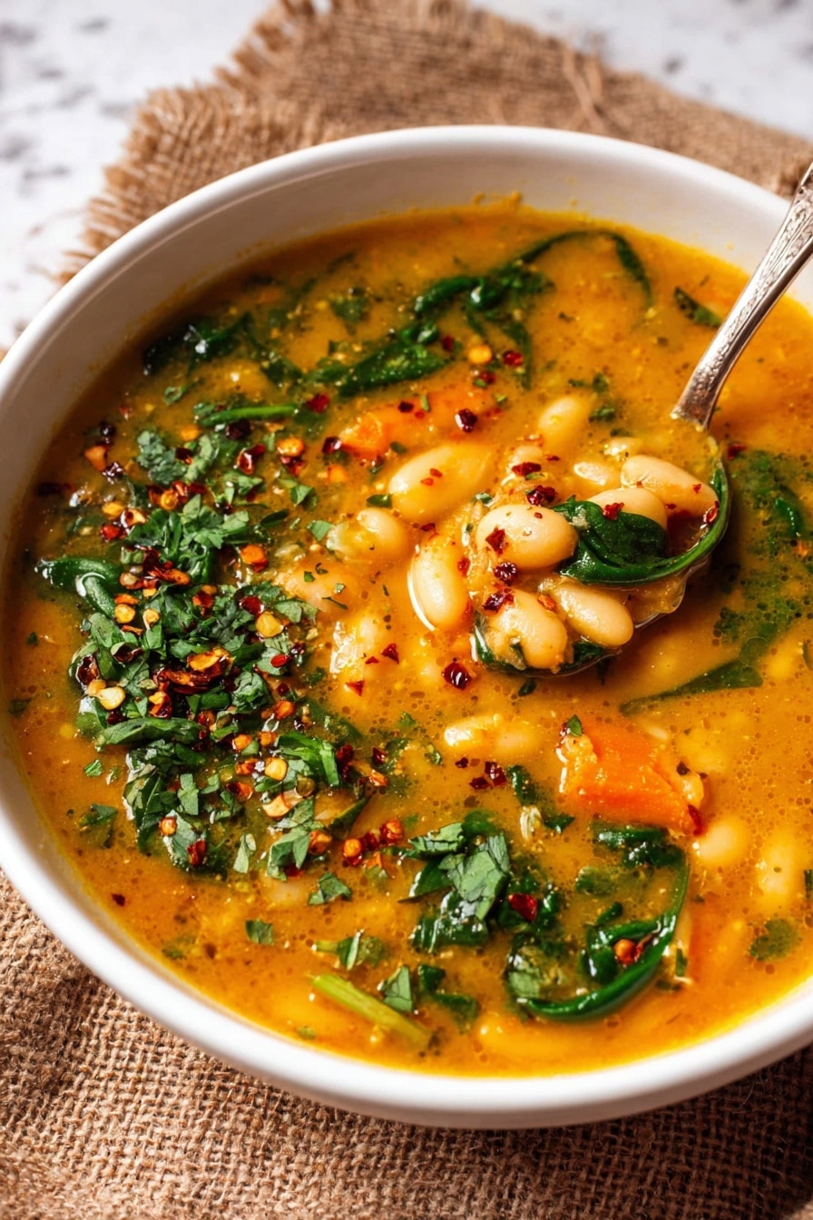 In a white bowl on a white marbled surface, there is a thick soup with a rich orange-yellow broth filled with white beans and small diced orange carrots. Bright green spinach leaves are mixed into the soup, adding fresh color. The soup is sprinkled with red chili flakes and small chopped green herbs on top, adding texture and contrast. A spoon inside the bowl holds a scoop of the soup, showing the beans, carrots, and bits of onions clearly. Photo taken with an iphone --ar 4:5 --v 7