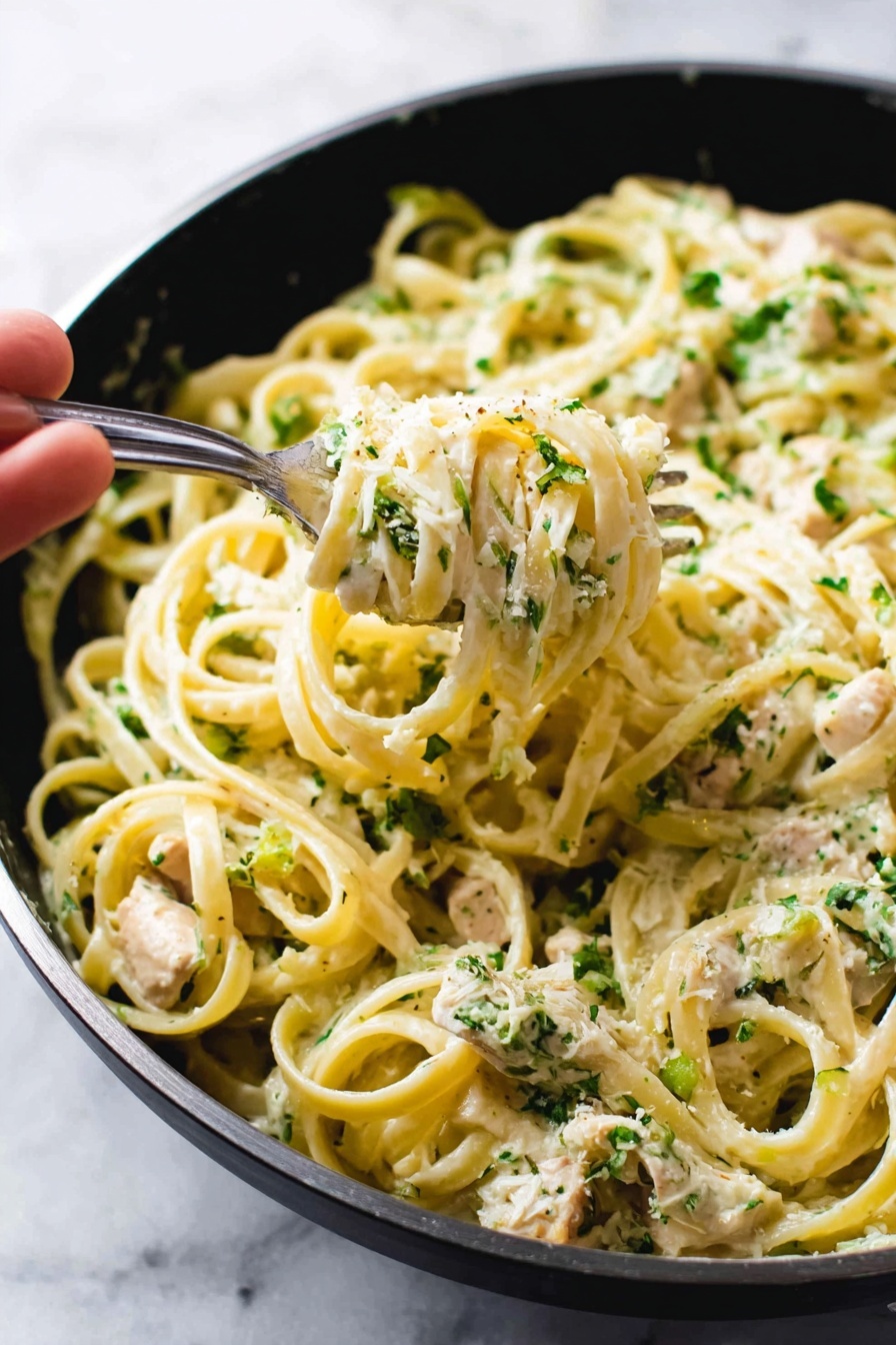 A white bowl holds a serving of fettuccine pasta mixed with small chunks of white cooked chicken and sprinkled with chopped green herbs, giving the dish a fresh and textured look. The pasta is light beige with a smooth texture, layered evenly with creamy sauce coating it. A metal fork rests inside the bowl, slightly buried in the pasta on the upper side. The bowl is placed on top of a soft gray cloth, all set on a white marbled surface photo taken with an iphone --ar 4:5 --v 7
