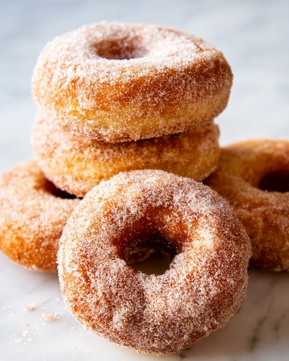 The image shows four round donuts with a rough texture, covered in a light brown layer of cinnamon sugar. One donut is broken in half, revealing a soft, pale yellow inside with a slightly crumbly texture. The donuts sit on a white marbled surface, with some sugar sprinkled around them. The overall look is warm and inviting, highlighting the contrast between the crunchy outside and soft inside of the donuts. photo taken with an iphone --ar 4:5 --v 7