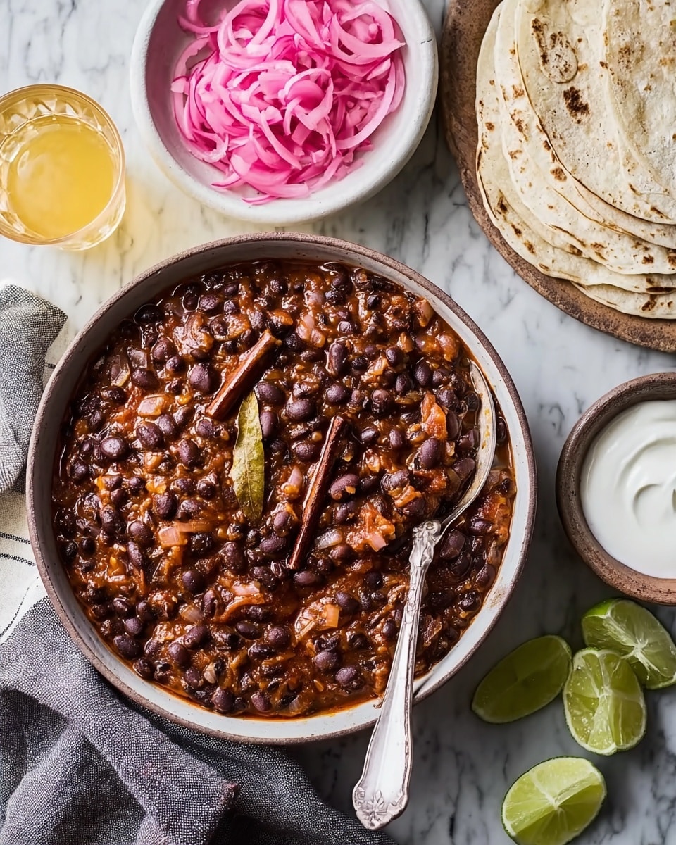 A large white bowl filled with a thick, chunky stew made of black beans, diced onions, and a rich reddish-brown sauce, topped with two bay leaves, and a cinnamon stick visible inside. There is a spoon resting in the bowl. On the right, there is a stack of three folded tortillas on a white plate, next to small lime wedges scattered on a white marbled texture surface. A small white bowl holds a smooth white sauce, with a spoon inside. Above, a white bowl contains bright pink pickled onions. Two clear glasses with a pale yellow liquid are placed on the white marbled surface, next to a beige and brown-striped cloth napkin. Photo taken with an iphone --ar 4:5 --v 7