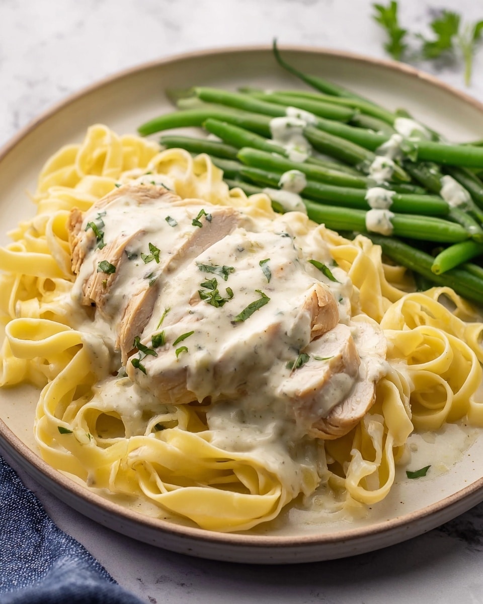 The dish shows three layers on a white plate sitting on a white marbled surface: the bottom layer is pale yellow fettuccine pasta with a soft texture, the middle layer is a piece of light beige chicken with a smooth surface, and the top layer is creamy white sauce with green herb specks evenly covering the chicken and a little on the pasta. On the side, there is a small pile of green beans with a slight sheen, also topped with the creamy white sauce. Some small green herb pieces are sprinkled across the dish, adding color contrast. Photo taken with an iphone --ar 4:5 --v 7