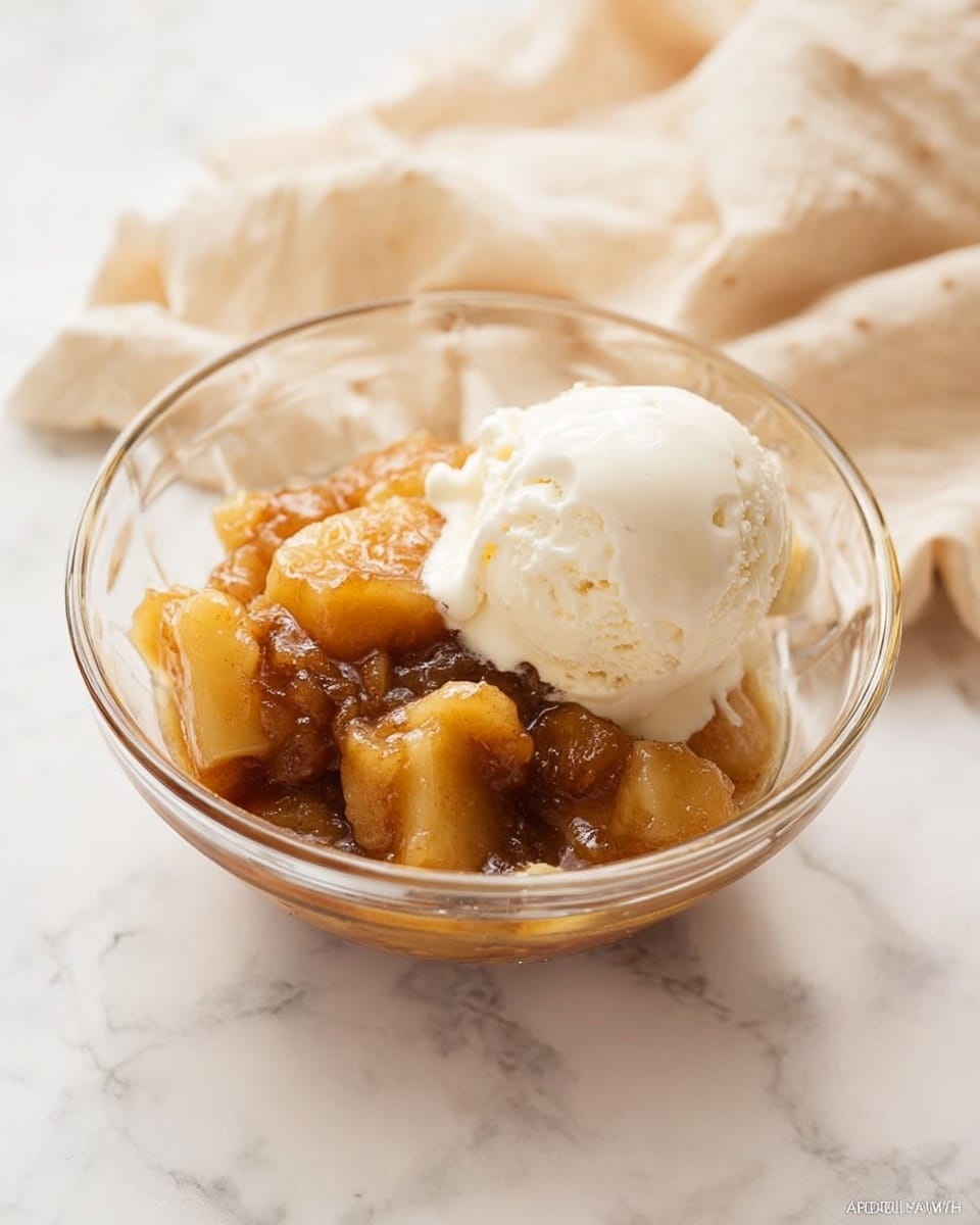 A clear glass bowl filled with two main layers: a bottom layer of soft, warm apple chunks mixed with a shiny, brown syrup, and on top, a large scoop of smooth, creamy vanilla ice cream that is starting to melt a little onto the warm apples. The bowl sits on a white marbled surface with a soft, beige cloth casually draped in the background. photo taken with an iphone --ar 4:5 --v 7