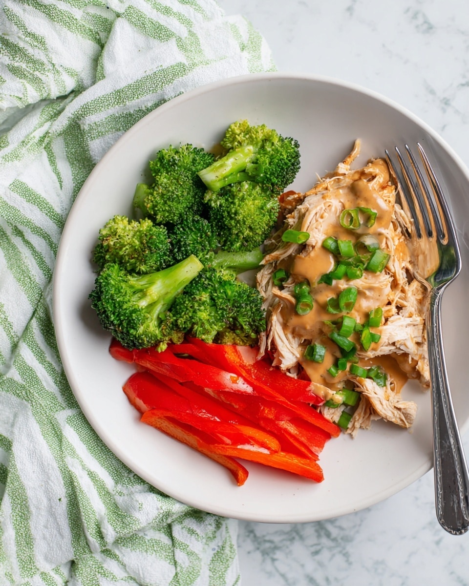 A white plate holds three main layers of food arranged side by side: bright green steamed broccoli florets on the left, vibrant red sliced bell peppers below them, and shredded light brown chicken topped with thick tan gravy and sprinkled chopped green onions on the right. A shiny silver fork rests on the right edge of the plate. The plate is set on a white marbled surface with a green and white striped cloth partially underneath. Photo taken with an iphone --ar 4:5 --v 7