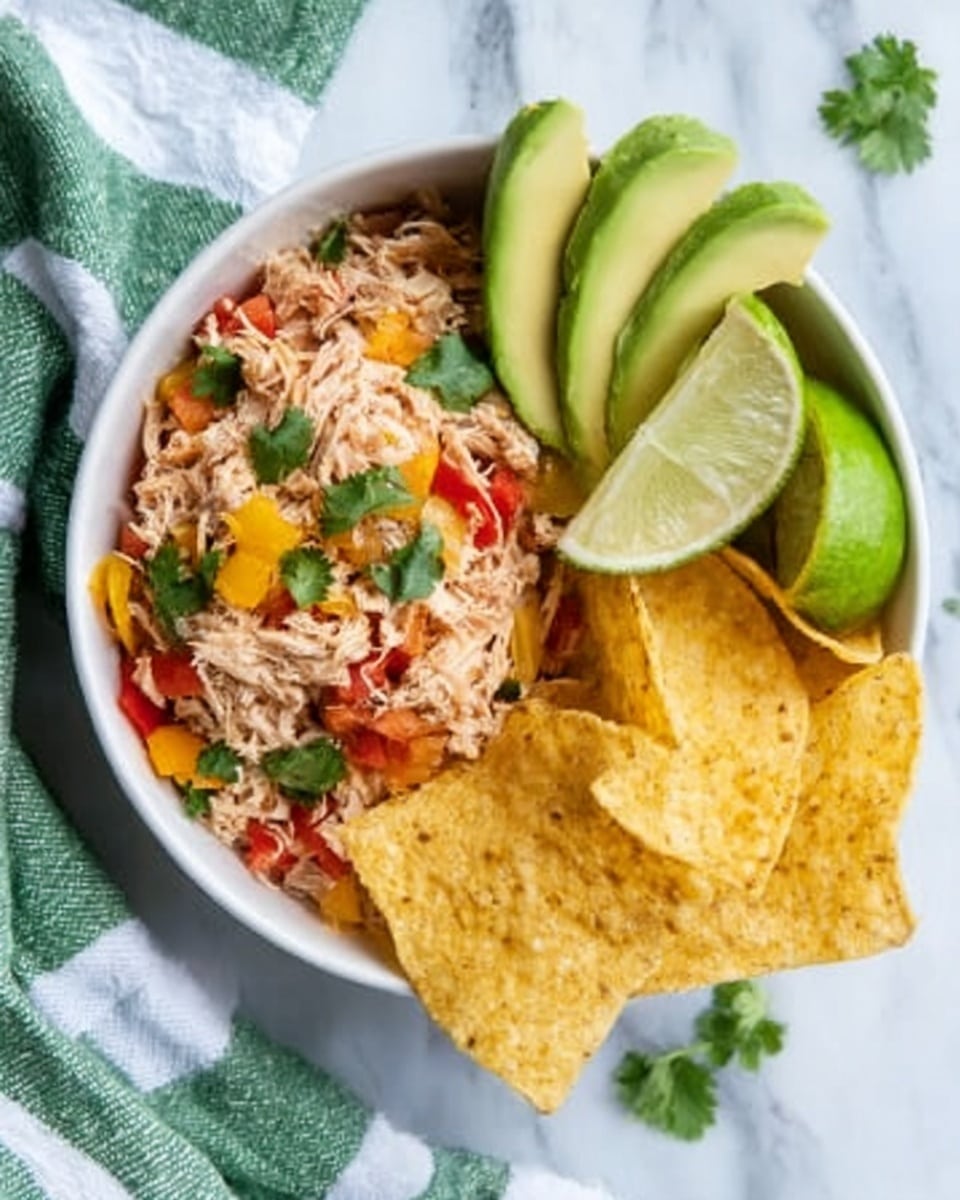 The image shows a white bowl filled with three main elements arranged in layers. On the left side, there is shredded chicken mixed with red and yellow bell peppers and small green cilantro leaves, creating a colorful, textured layer. Next to it, on the right side of the bowl, are several golden-yellow tortilla chips with a rough, crispy texture, stacked loosely. On top of the bowl’s right edge, there are three thin slices of light green avocado neatly placed in a fan shape. A wedge of lime is tucked close to the shredded chicken, adding a slight bright green contrast. The bowl sits on a white marbled surface with a green and white striped cloth partially visible nearby. The photo taken with an iphone --ar 4:5 --v 7