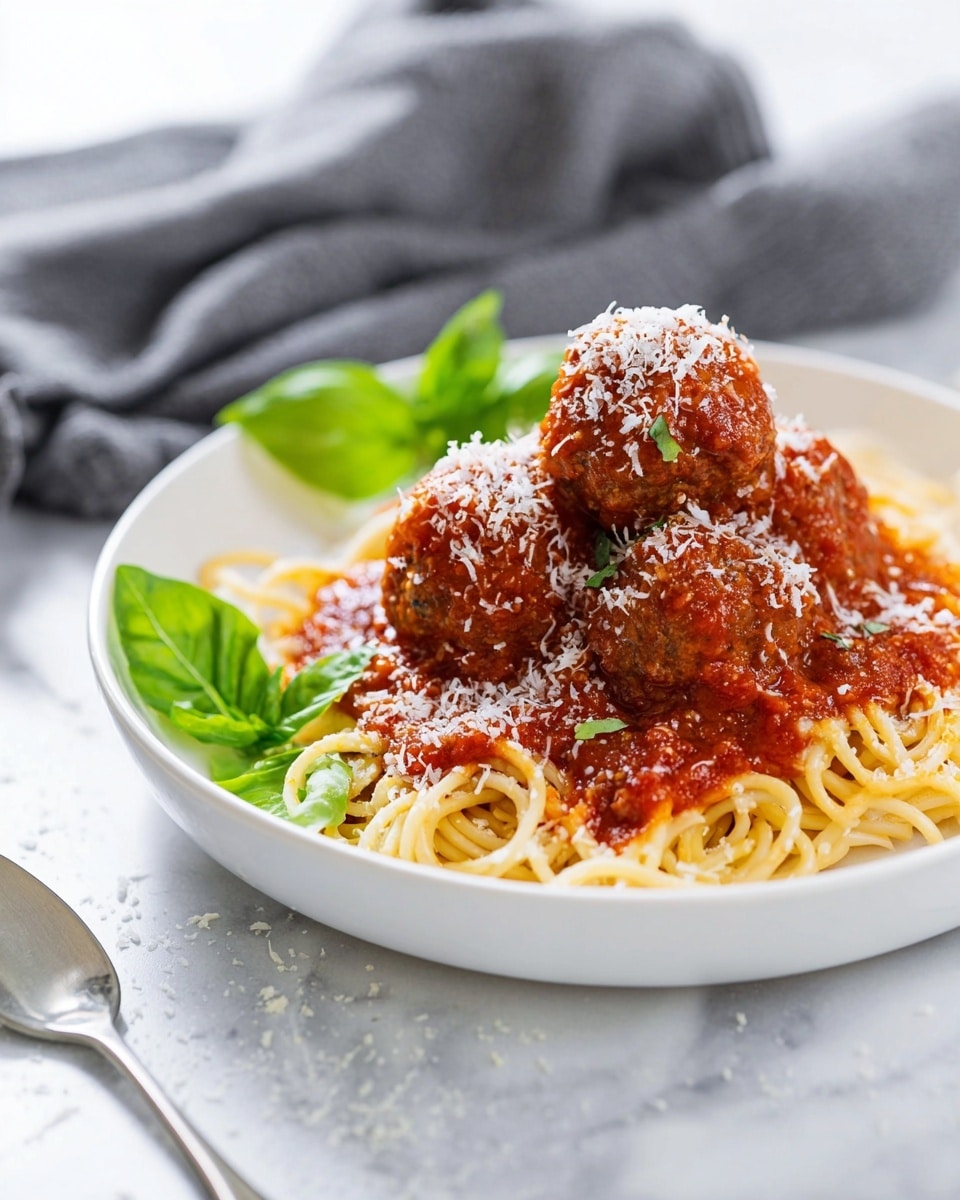 A white plate holds a serving of twisted spaghetti noodles at the bottom, topped with four round meatballs covered in thick, red tomato sauce. The meatballs are sprinkled with white grated cheese. Two fresh green basil leaves rest on the left side of the meatballs for garnish. The plate is set on a white marbled surface with a spoon placed in front and a gray cloth blurred in the background. Photo taken with an iphone --ar 4:5 --v 7