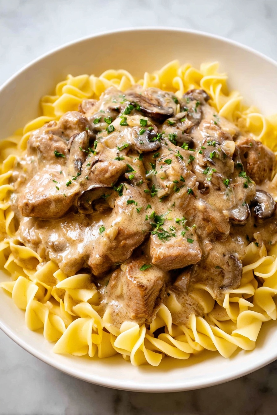 A white bowl with a bed of yellow egg noodles arranged in a ring around the edges, with a thick layer of creamy light brown sauce covered with cooked mushrooms and strips of meat in the center. The sauce is smooth with small green herb pieces sprinkled on top. The whole dish is placed on a white marbled surface. photo taken with an iphone --ar 4:5 --v 7