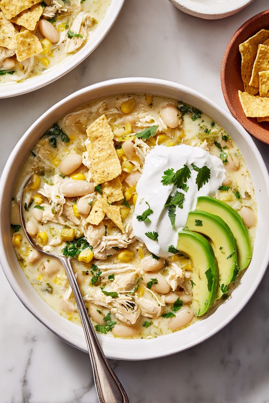 A white bowl filled with creamy chicken soup containing visible shredded chicken pieces, white beans, corn kernels, and chopped green herbs mixed throughout the light broth. On the surface, there are broken light yellow tortilla chips scattered. On one side of the bowl, there is a neat layer of three bright green avocado slices arranged overlapping each other. At the center top, a round dollop of smooth white sour cream topped with a few small green herb leaves adds contrast. A silver spoon rests inside the bowl on the left edge. The bowl sits on a white marbled surface with part of another bowl and an orange terracotta plate with tortilla chips visible at the edges. Photo taken with an iphone --ar 4:5 --v 7
