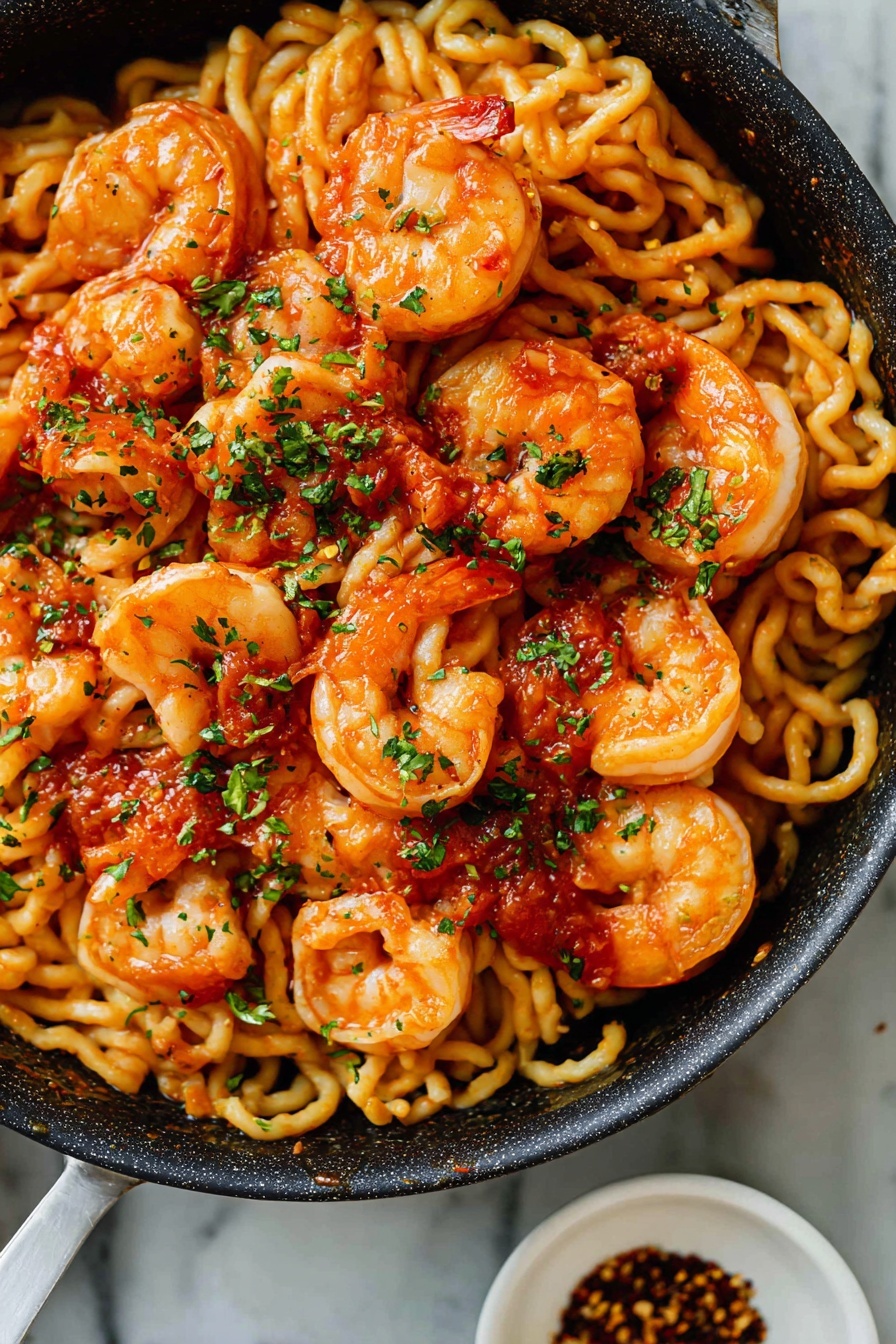 A close-up of a black pan filled with a shrimp pasta dish featuring curly, light yellow pasta noodles as the base layer. On top, there are plump, orange-pink cooked shrimp scattered evenly across the noodles. Bright red tomato sauce is dotted throughout the dish in small patches, adding color contrast. The dish is sprinkled with finely chopped green herbs and black pepper flakes for texture and garnish. The pan sits on a white marbled surface, and in the corner, a small white bowl with dark sauce and red chili flakes is partially visible. Photo taken with an iphone --ar 4:5 --v 7