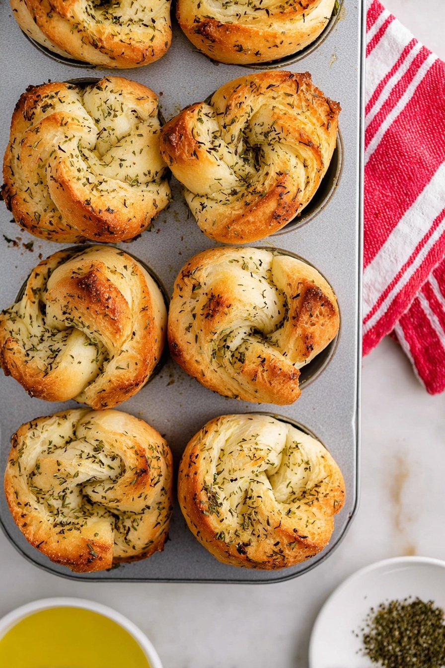 The image shows a close-up of six golden brown herb rolls in a silver muffin tray, each roll having multiple soft, airy layers of dough speckled with green dried herbs, giving a slightly rough texture. The rolls sit in each circular muffin cup, with some edges nicely browned and slightly crisp. The silver tray is on a white marbled surface with a red and white striped cloth at the top right corner. The lighting softly highlights the warm tones and texture of the rolls. photo taken with an iphone --ar 4:5 --v 7