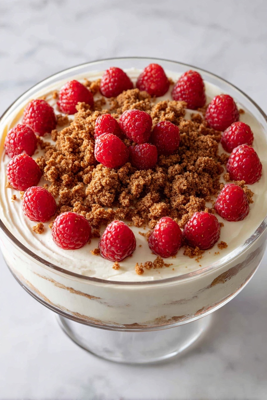The image shows a clear glass trifle bowl with three main layers. The bottom layer is dark brown cake pieces, followed by a smooth pale yellow custard layer, then a white creamy layer sprinkled lightly with brown crumbs. This pattern repeats once more, topped with a final creamy white layer covered with a ring of brown crumbs and a cluster of bright red raspberries in the center. The bowl stands on a base with a pink ribbon underneath and is placed on a white marbled surface with a blurred green pine background. Photo taken with an iphone --ar 4:5 --v 7