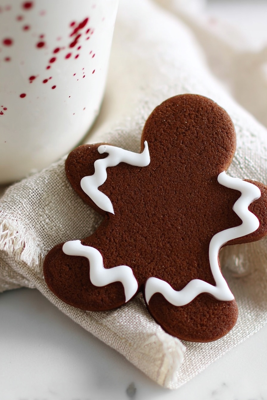 The image shows a single gingerbread cookie shaped like a star with five points. The cookie is dark brown with a slightly rough texture. Around the edge of the cookie, there is a thick white icing line that follows the shape closely with soft curves. One point of the cookie has a small bite taken out of it, revealing a slightly lighter brown inside. The cookie sits on a cream textured cloth that is folded over a white marbled surface. Part of a white ceramic cup is visible behind the cookie, but mostly out of focus. The lighting is soft and natural, casting gentle shadows. photo taken with an iphone --ar 4:5 --v 7