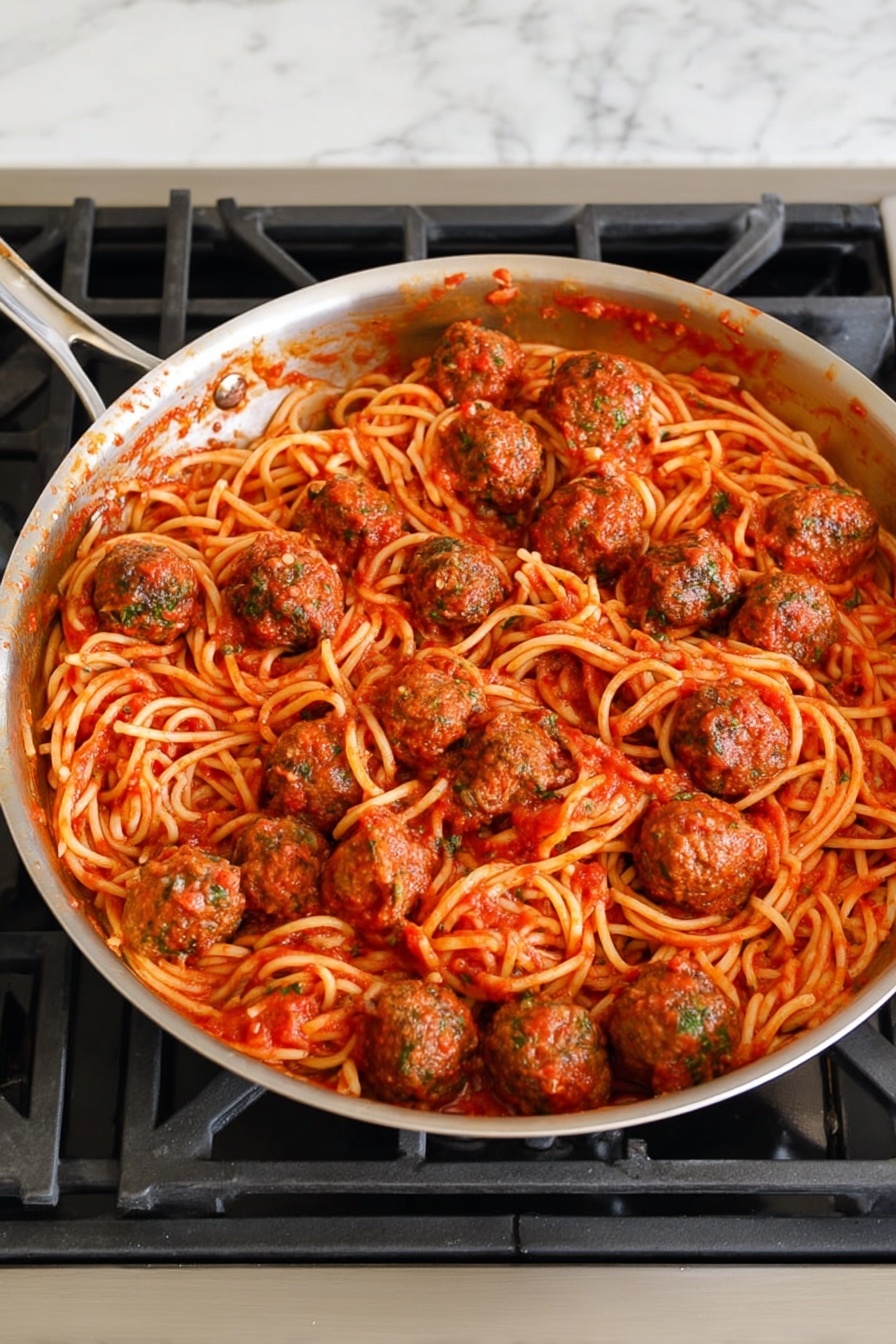 A silver frying pan filled with one layer of spaghetti noodles covered in a red tomato sauce, mixed with about twelve round meatballs evenly spread on top, each meatball showing a textured surface with bits of green herbs. The pan sits on a gas stove with black grates, and the surrounding surface is a white marbled texture. photo taken with an iphone --ar 4:5 --v 7