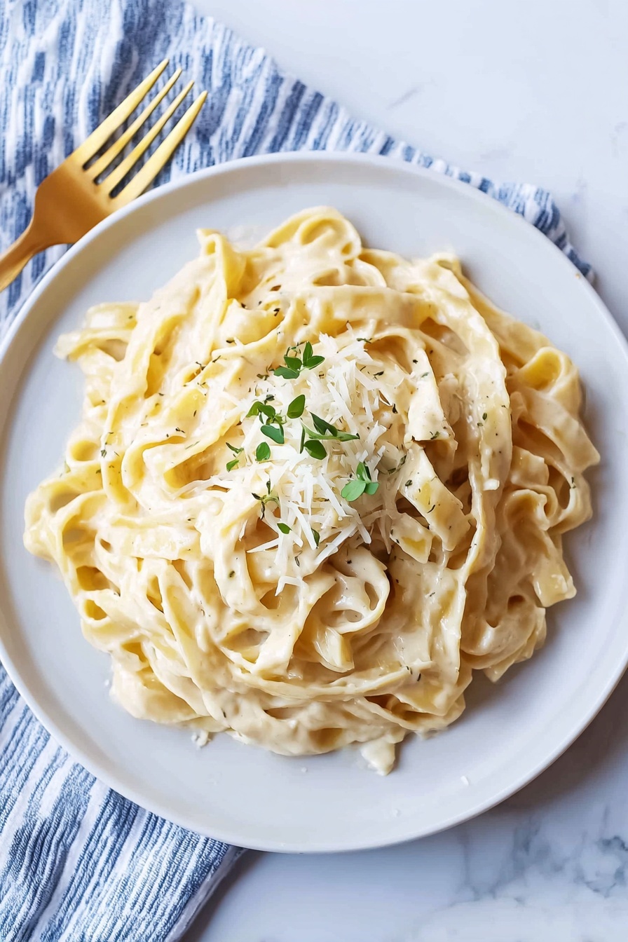 A white plate filled with creamy fettuccine pasta, layered with smooth, thick Alfredo sauce giving the pasta a shiny, light beige color. On top, there is a small heap of finely grated cheese that looks slightly crumbly and pale yellow, sprinkled with small pieces of chopped green herbs for color contrast. The pasta strands are wide and curled in a loose heap. The plate is placed on a white marbled surface beside a blue and white striped cloth napkin with a gold fork resting on it. Photo taken with an iphone --ar 4:5 --v 7