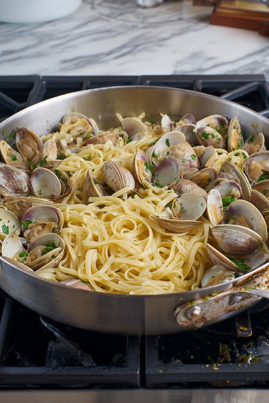 The image shows a large silver pan filled with cooked fettuccine pasta mixed with open clams. The fettuccine is pale yellow and looks soft with some glistening from light oil or butter. The clams have shells in shades of light brown, beige, and white, scattered evenly throughout the pasta. There are small pieces of green herbs sprinkled on top, adding a fresh touch. The pan sits on a stove with black grates, and the surrounding surface has a white marbled texture. photo taken with an iphone --ar 4:5 --v 7