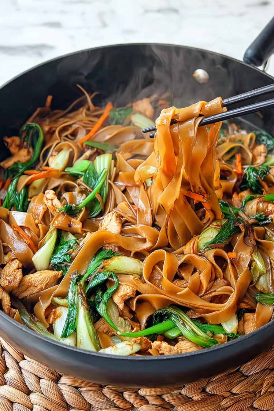A black pan filled with stir-fried wide flat brown noodles mixed with light brown cooked chicken pieces and bright green leafy vegetables with crispy stems, thin orange carrot strips are also mixed inside; a pair of black chopsticks is lifting some noodles in the center, showing the shiny and soft texture of the noodles and the colorful mix of ingredients underneath, steam rises gently from the dish, and the pan rests on a beige woven mat over a white marbled surface photo taken with an iphone --ar 4:5 --v 7