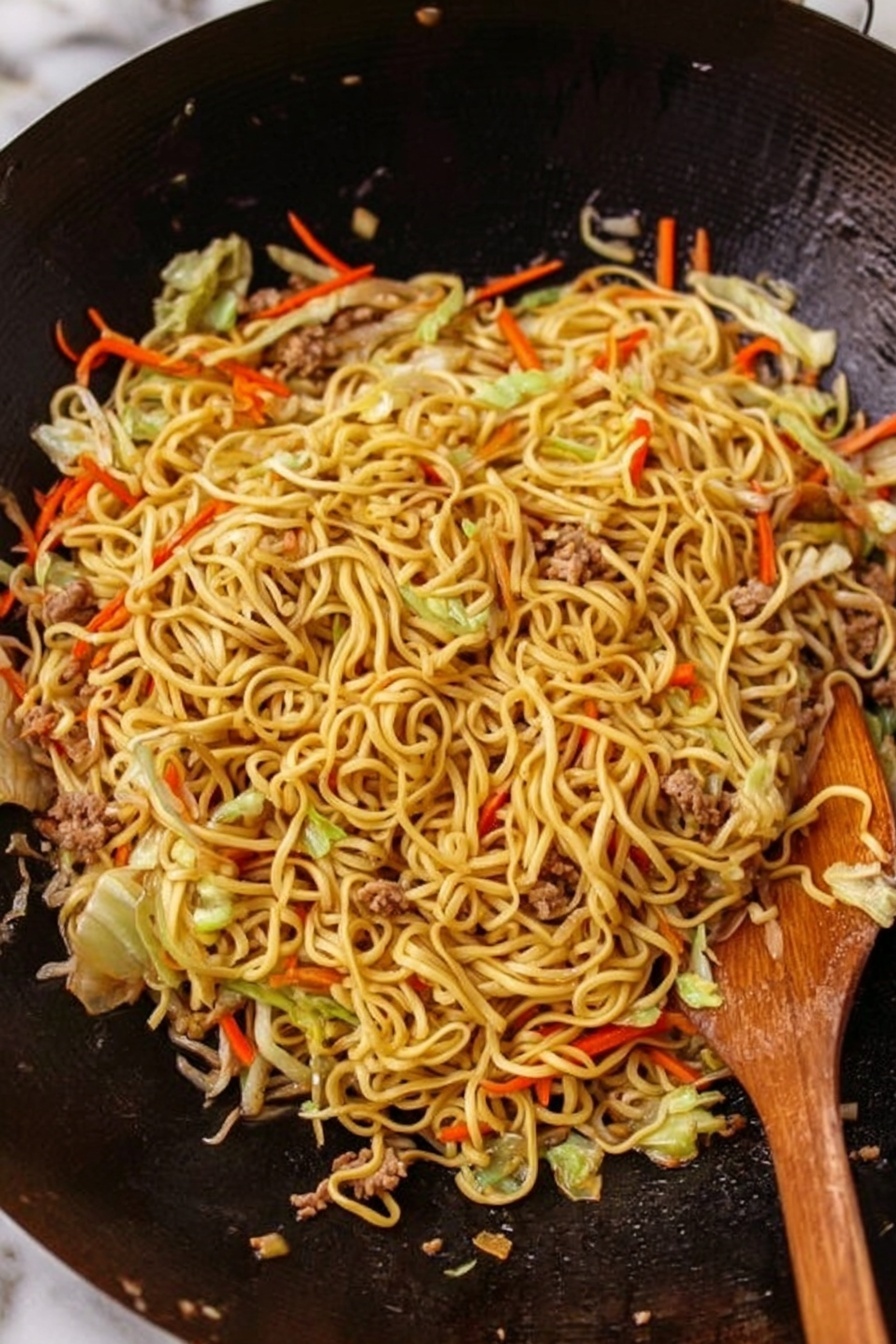 The image shows a large black wok filled with stir-fried noodles. The noodles are light brown and glossy, mixed with thin strips of orange carrots, white cabbage, and green vegetables. Small pieces of cooked meat are scattered throughout, adding texture. The noodles and vegetables are evenly spread across the wok, with some wooden chopsticks resting on the right side. The background is a white marbled surface. Photo taken with an iphone --ar 4:5 --v 7