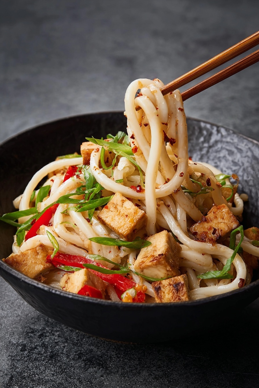 A bowl of thick, creamy noodles tangled with red pepper strips and small golden brown tofu cubes sits on a dark surface replaced with a white marbled texture. The dish is topped with thin bright green spirals of scallions and scattered chopped peanuts, all mixed with a light sauce giving a slight shine to the noodles. The tofu cubes show light browning and the entire bowl is black, creating contrast with the food colors photo taken with an iphone --ar 4:5 --v 7