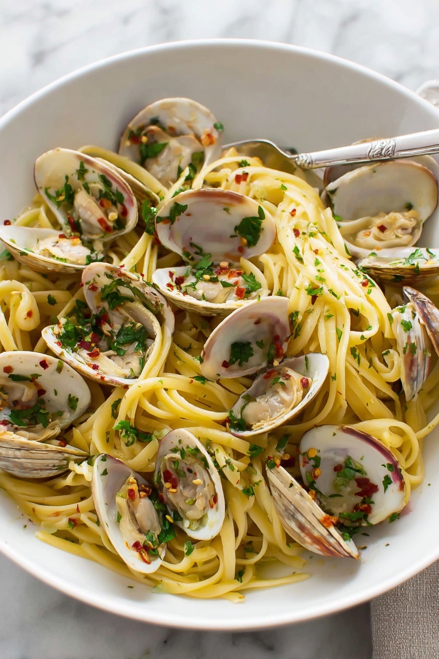 A white bowl holds a serving of spaghetti tangled in the center with a light yellow color, mixed with open clam shells showing pale beige clam meat inside. The clams are scattered evenly on top and around the pasta, garnished with small chopped green herbs and flecks of red chili flakes. The dish looks moist with hints of a light sauce and small bits of white garlic pieces. A silver fork is placed resting on the right side inside the bowl. The bowl is set on a white marbled surface with a glass of water blurred in the background. Photo taken with an iphone --ar 4:5 --v 7