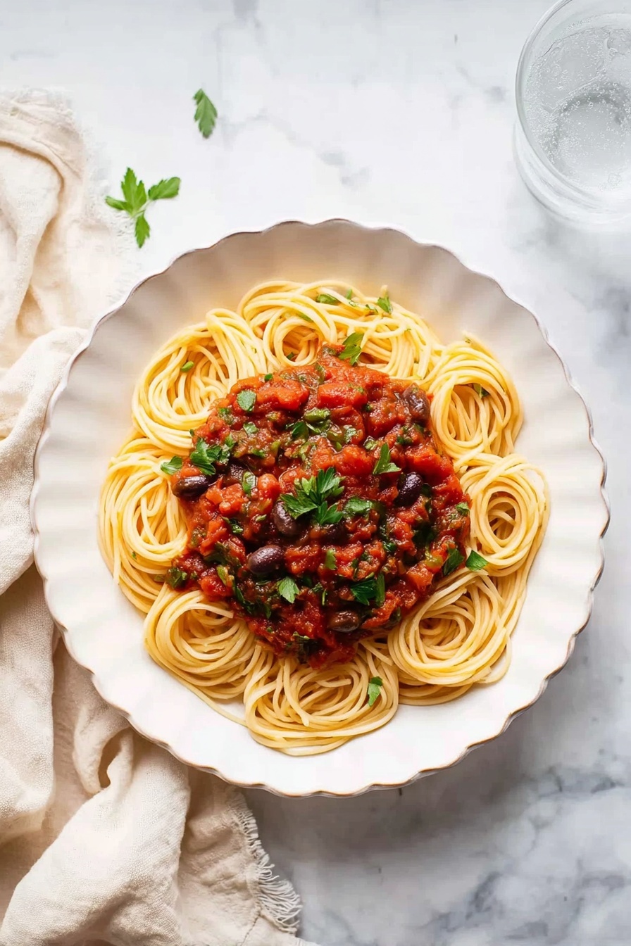 A white plate filled with spaghetti layered neatly in the center, topped with a chunky red tomato sauce mixed with pieces of dark olives and sprinkled green parsley leaves. The sauce sits mostly on top and around the pasta, giving a mix of red, green, and pale beige colors. A silver fork rests on the edge of the plate, partly twirling some of the spaghetti. The plate is placed on a white marbled surface with a glass of clear water at the top right and a beige cloth to the left. photo taken with an iphone --ar 4:5 --v 7