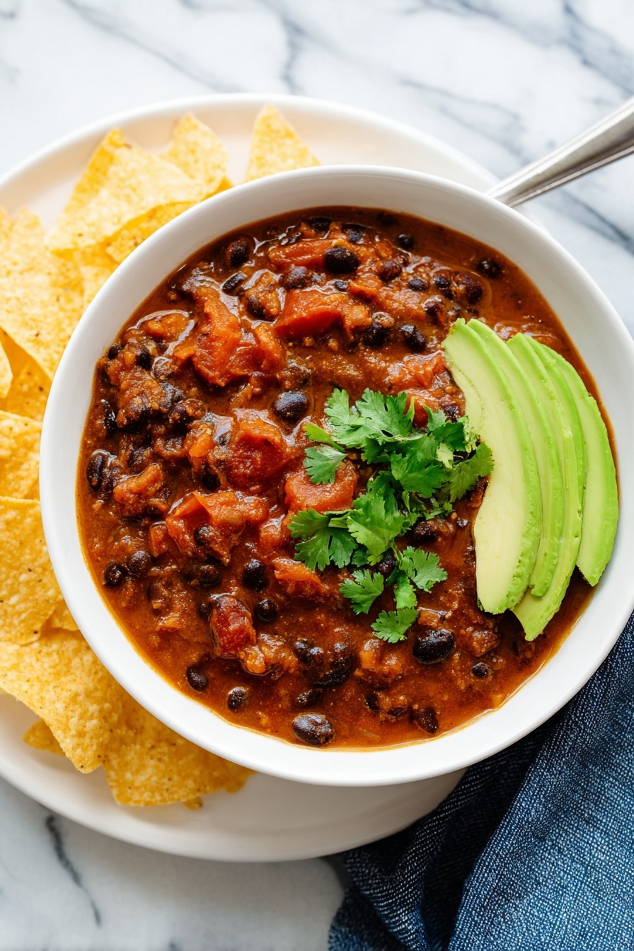 A white bowl filled with thick chili showing black beans, chunks of red tomatoes, and pieces of vegetables in a rich reddish-brown sauce, topped with fresh small green cilantro leaves. On one side, several slices of bright green avocado are neatly arranged. The bowl sits on a white plate with a few light yellow triangular tortilla chips placed on the plate's left side. The whole dish is on a white marbled surface with a blue cloth partially visible on the lower right side. Photo taken with an iphone --ar 4:5 --v 7