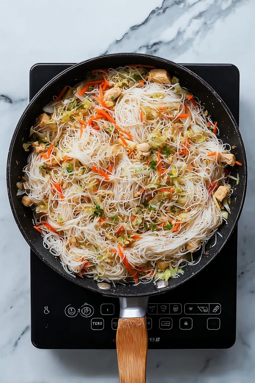 A black pan filled with a mix of thin white noodles, orange carrot strips, light green cabbage pieces, and small chunks of cooked light brown chicken, all lightly mixed together. The pan has a wooden handle and sits on a black electric cooker, all set on a white marbled surface. The colors contrast well, showing the soft texture of the noodles, the crispness of the vegetables, and the tender cooked chicken. Photo taken with an iphone --ar 4:5 --v 7