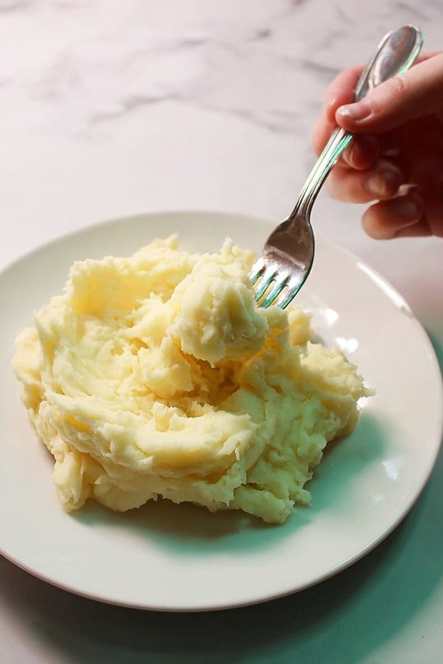 The image shows a white bowl filled with smooth, creamy mashed potatoes that have a pale yellow color with small specks of seasoning. A spoon rests inside the bowl, partially scooping out some of the soft potatoes, revealing a light, fluffy texture underneath the slightly textured top layer. The bowl sits on a white marbled surface, adding a clean and simple background to the warm, comforting dish. photo taken with an iphone --ar 4:5 --v 7