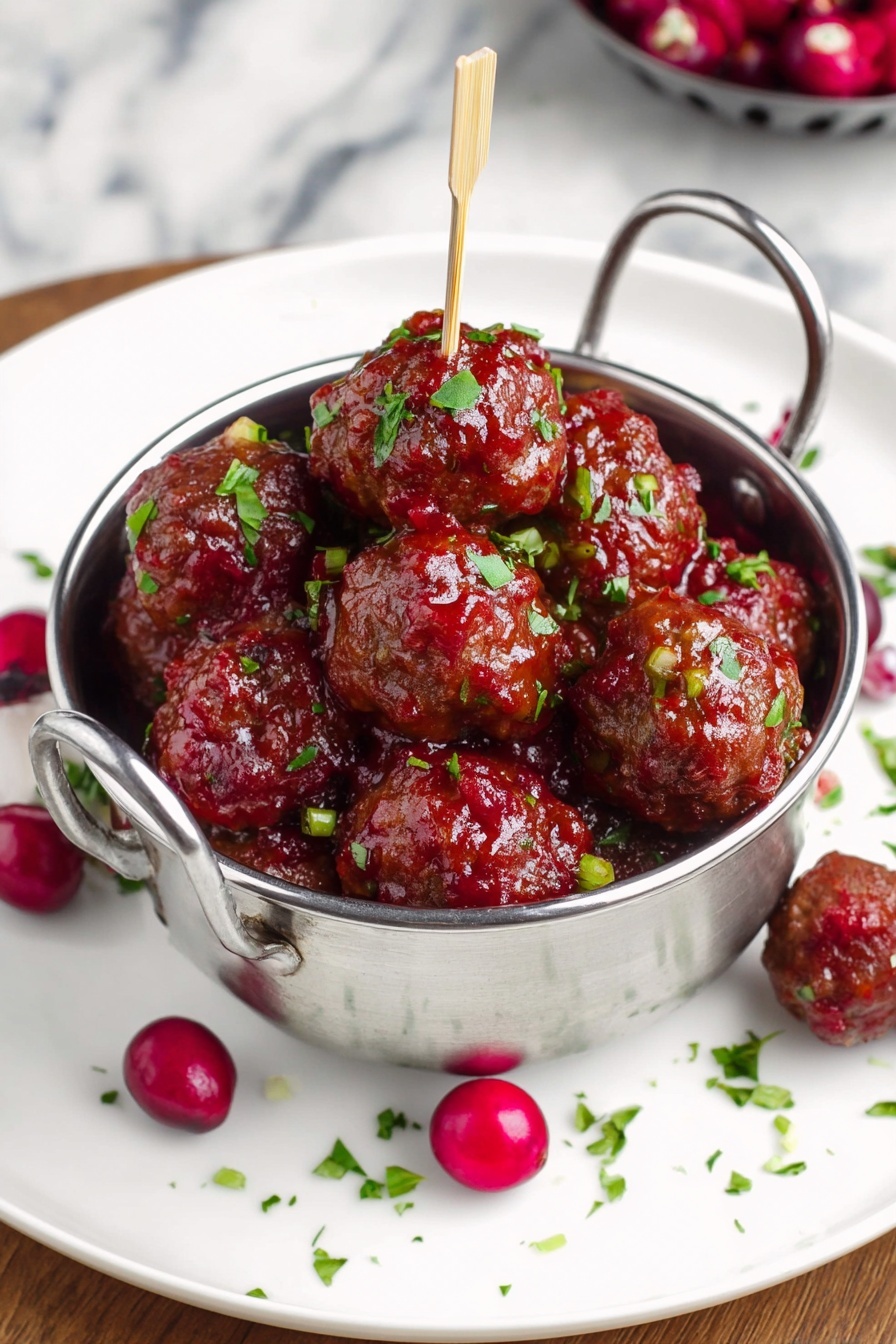 A round metal bowl with two handles holds many brown meatballs covered in a shiny red sauce, scattered with small green herb pieces and a few bright red whole cranberries. The bowl sits on a white plate with a few more cranberries and green herbs scattered around it. One meatball is on a wooden toothpick resting on the edge of the plate. The whole scene is set on a white marbled surface with a clear glass bowl of green herbs partially visible in the top right corner. photo taken with an iphone --ar 4:5 --v 7