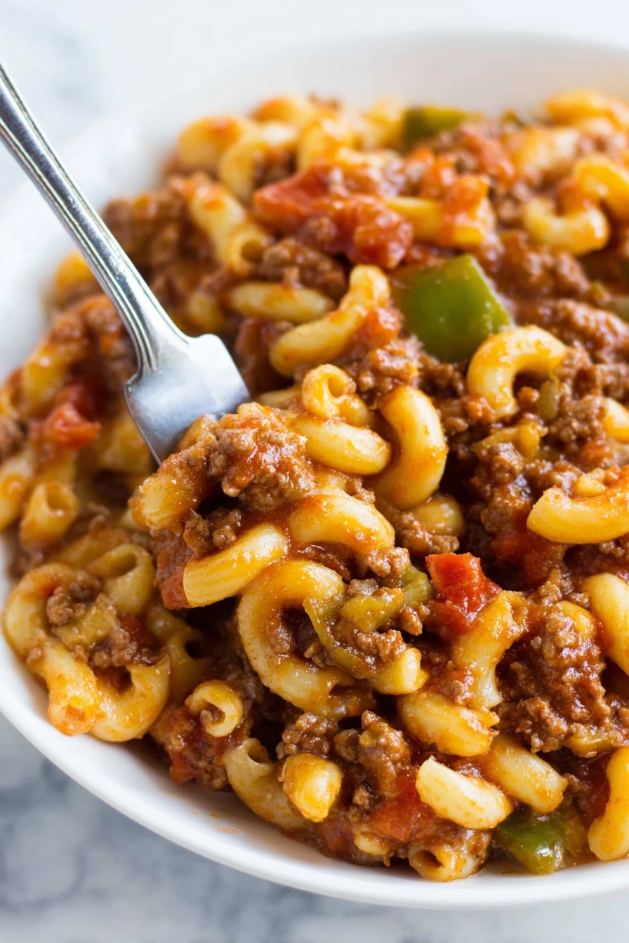 The image shows a close-up of a white bowl filled with a rich mixture of ground meat and elbow macaroni pasta. The pasta is light yellow and smooth, coated in a thick reddish-brown sauce that also covers the browned, crumbly meat. There are visible small pieces of green bell pepper and bits of tomato mixed evenly throughout. A knife is partially inserted into the bowl, ready to scoop some of the hearty pasta dish. The background surface is a white marbled texture. photo taken with an iphone --ar 4:5 --v 7
