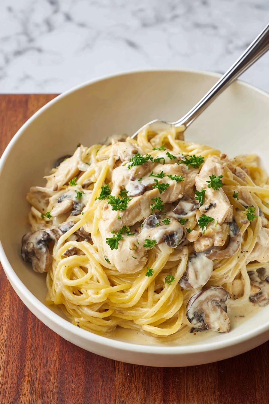 A white bowl holds a creamy pasta dish with about two layers visible: the base layer is a sauce mixed with pieces of pale chicken and brown mushrooms, all coated in a glossy, light cream sauce. On top, there is a layer of yellowish spaghetti noodles, some twisted and resting loosely, garnished with small bright green parsley leaves. A silver fork leans against the side of the bowl, partially submerged in the dish. The bowl sits on a white marbled surface. photo taken with an iphone --ar 4:5 --v 7
