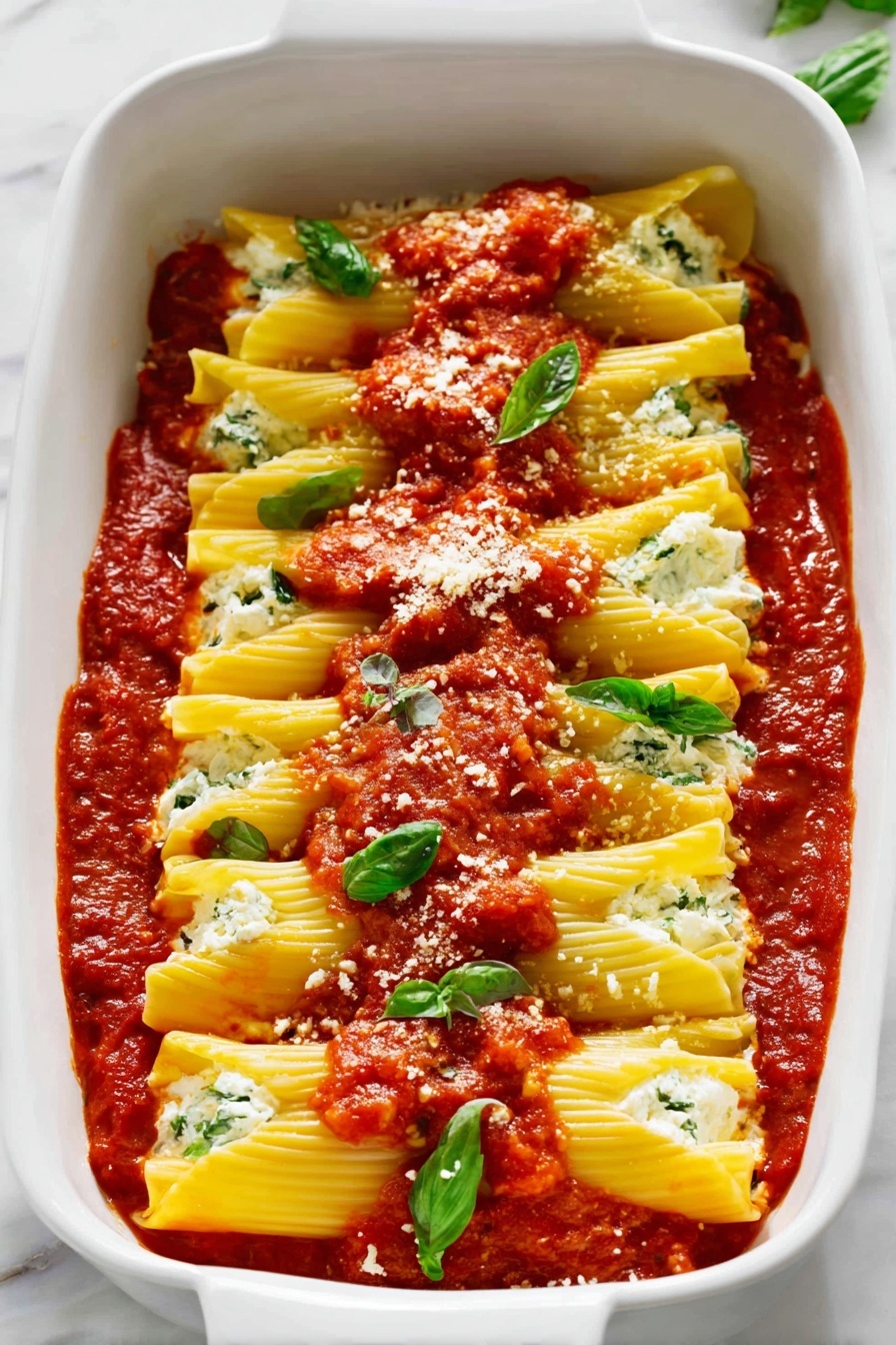 The image shows a white baking dish filled with layered pasta rolls standing side by side, each pale yellow pasta sheet slightly curved and stuffed with a creamy white filling with small green bits. On top of the pasta rolls is a thick layer of bright red tomato sauce, unevenly spread, with fresh green basil leaves and small grated white cheese sprinkled over it. The tomato sauce surrounds the pasta rolls at the bottom of the dish, creating a rich red base that contrasts with the pale pasta and white filling. The whole dish rests on a white marbled surface photo taken with an iphone --ar 4:5 --v 7