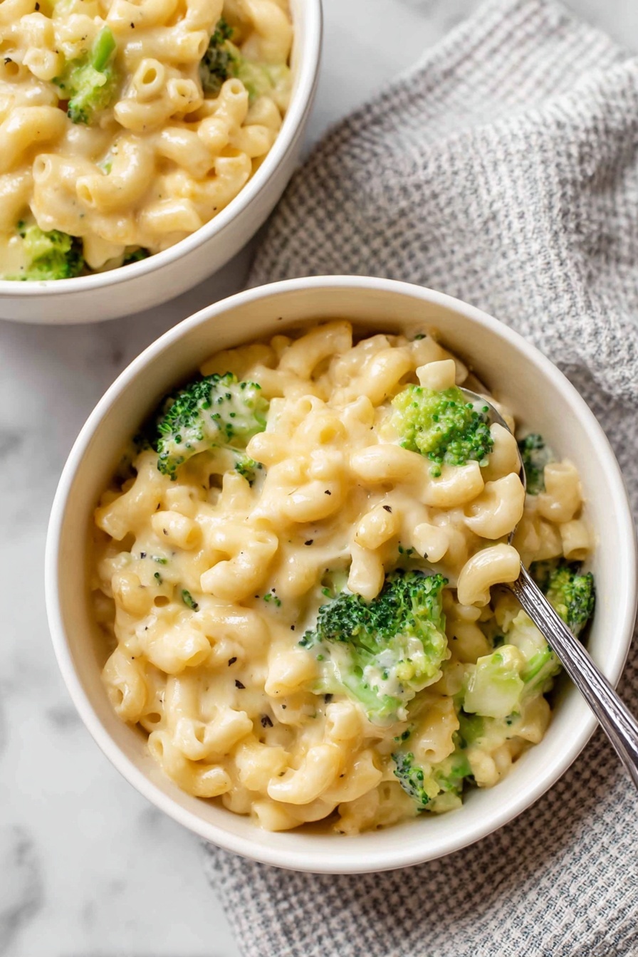 Two white bowls filled with creamy macaroni and cheese mixed with bright green broccoli florets. The pasta is coated in a smooth, light yellow cheese sauce with a glossy texture. Each bowl shows a good mix of pasta and vibrant broccoli pieces on top and throughout. The bowls sit on a white marbled surface. Nearby, there is a wrapped block of butter with a brown and blue label sitting on a white cloth with black stripes. Some uncooked elbow macaroni pieces are scattered loosely on the surface. Photo taken with an iphone --ar 4:5 --v 7