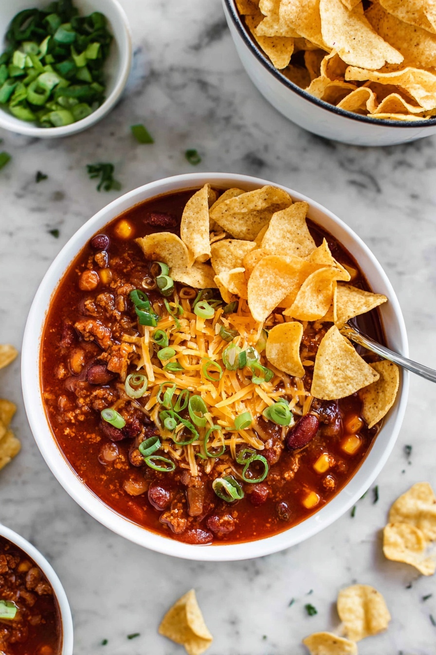 A white bowl filled with thick, dark red chili that contains visible kidney beans and small pieces of meat, topped with light yellow shredded cheese that is slightly melted into the chili. On top of the cheese, there are lightly golden corn chips arranged in a small pile, garnished with chopped green onions scattered on the chips and around the bowl's edge. The bowl sits on a white marbled surface, with a close-up view showing the rich texture of the chili, and a blurred second bowl of corn chips in the background. photo taken with an iphone --ar 4:5 --v 7