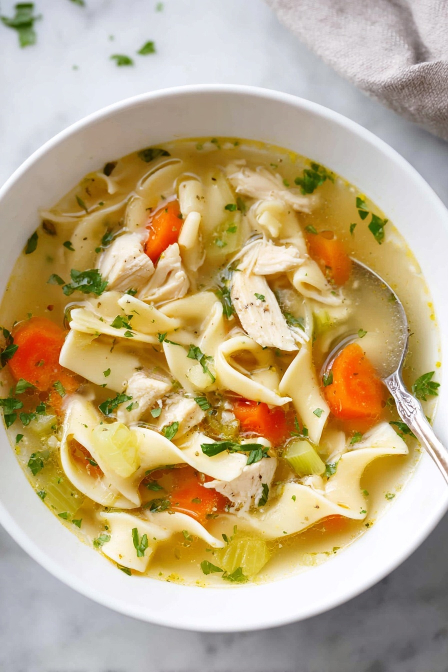 The image shows a white bowl filled with chicken noodle soup placed on a white marbled surface. The soup has a clear, yellowish broth with small green herbs sprinkled on top. Inside the soup, there are several chunky pieces of white chicken meat, some flat, wide egg noodles that curl softly, slices of orange carrot rounds, and light green celery slices. A shiny silver spoon sits next to the bowl, and there is a woman's hand slightly visible at the edge, as if about to hold the bowl. The texture of the soup looks smooth and warm, with a fresh, homemade feel. Photo taken with an iphone --ar 4:5 --v 7