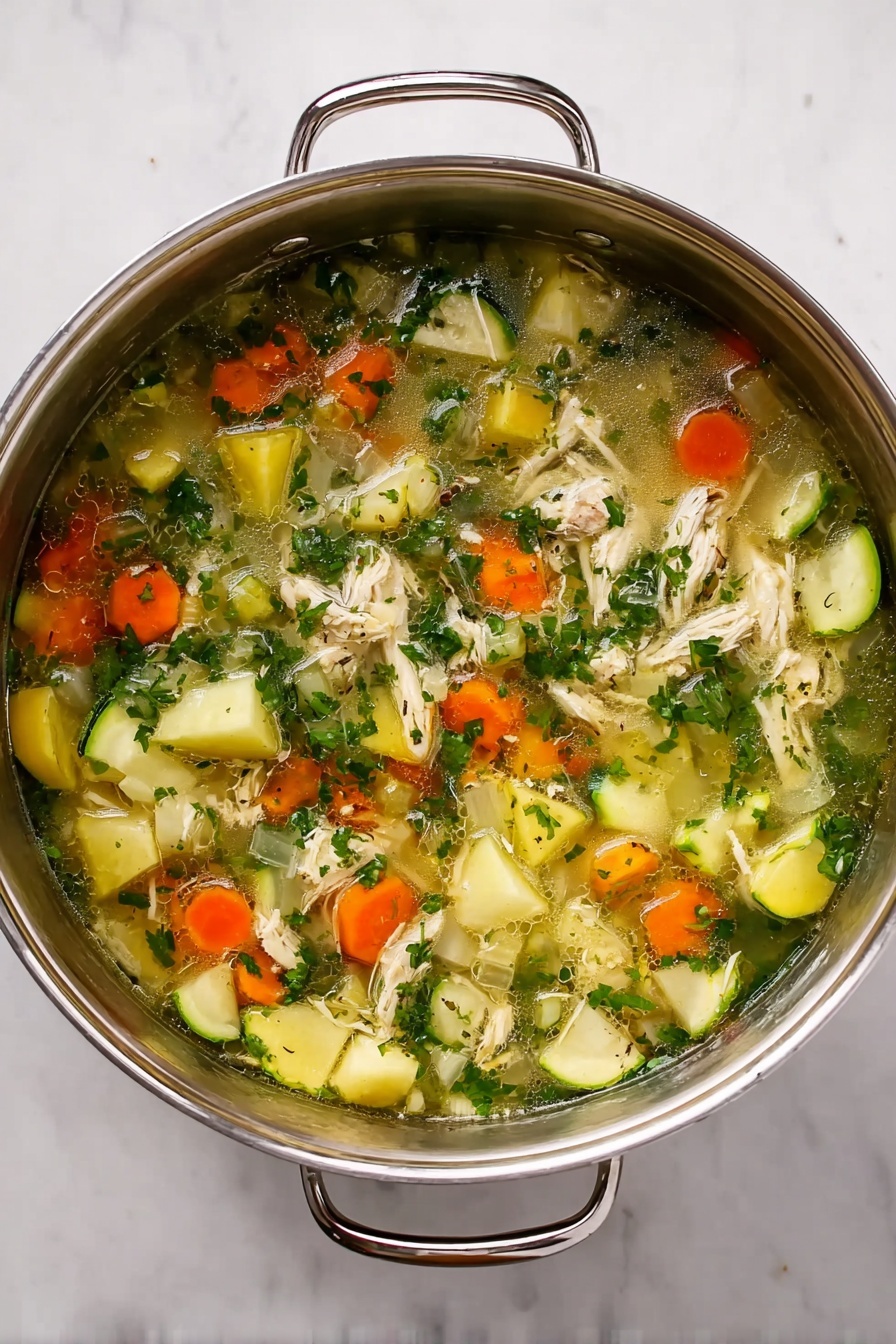 A silver pot filled with a colorful soup rests on a white marbled surface. The soup has a clear broth base with small pieces of shredded white meat and large chunks of soft vegetables. The vegetables include bright orange cubes, pale yellow potato pieces, translucent onion chunks, and green herbs mixed throughout. The soup looks fresh, with a mix of smooth and chunky textures visible. The pot has two handles, one on each side. photo taken with an iphone --ar 4:5 --v 7
