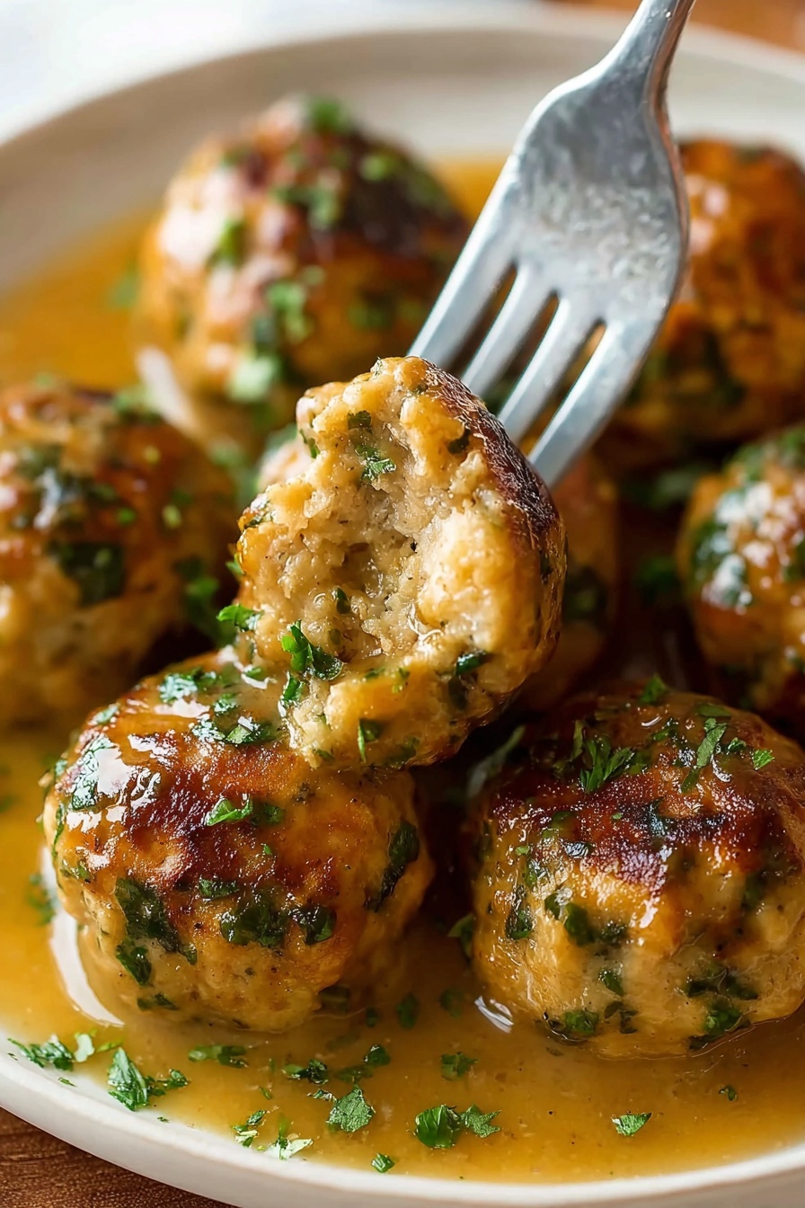 The image shows several round meatballs with a golden-brown crust and green herb bits scattered through them, resting in a light brown gravy. A silver fork holds one meatball with a bite taken out, showing a soft and moist inside with visible herbs. The meatballs are arranged closely on a white plate with some chopped green herbs sprinkled on top and the gravy pooling around them. The background is a white marbled texture photo taken with an iphone --ar 2:3 --v 7 - Stuffing Balls, Stuffing Balls Recipe, Easy Stuffing Balls, Holiday Side Dishes, Comfort Food Recipes